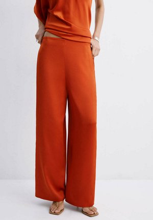 Mujer con pantalones de tiro alto, de pernera ancha en color naranja quemado y top sin mangas a juego, de pie descalza sobre un suelo claro.