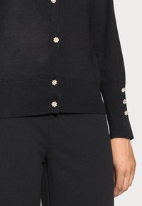 Black knitted cardigan with pearl-like buttons down the front and on the sleeve cuffs. Features a ribbed hem and smooth texture.