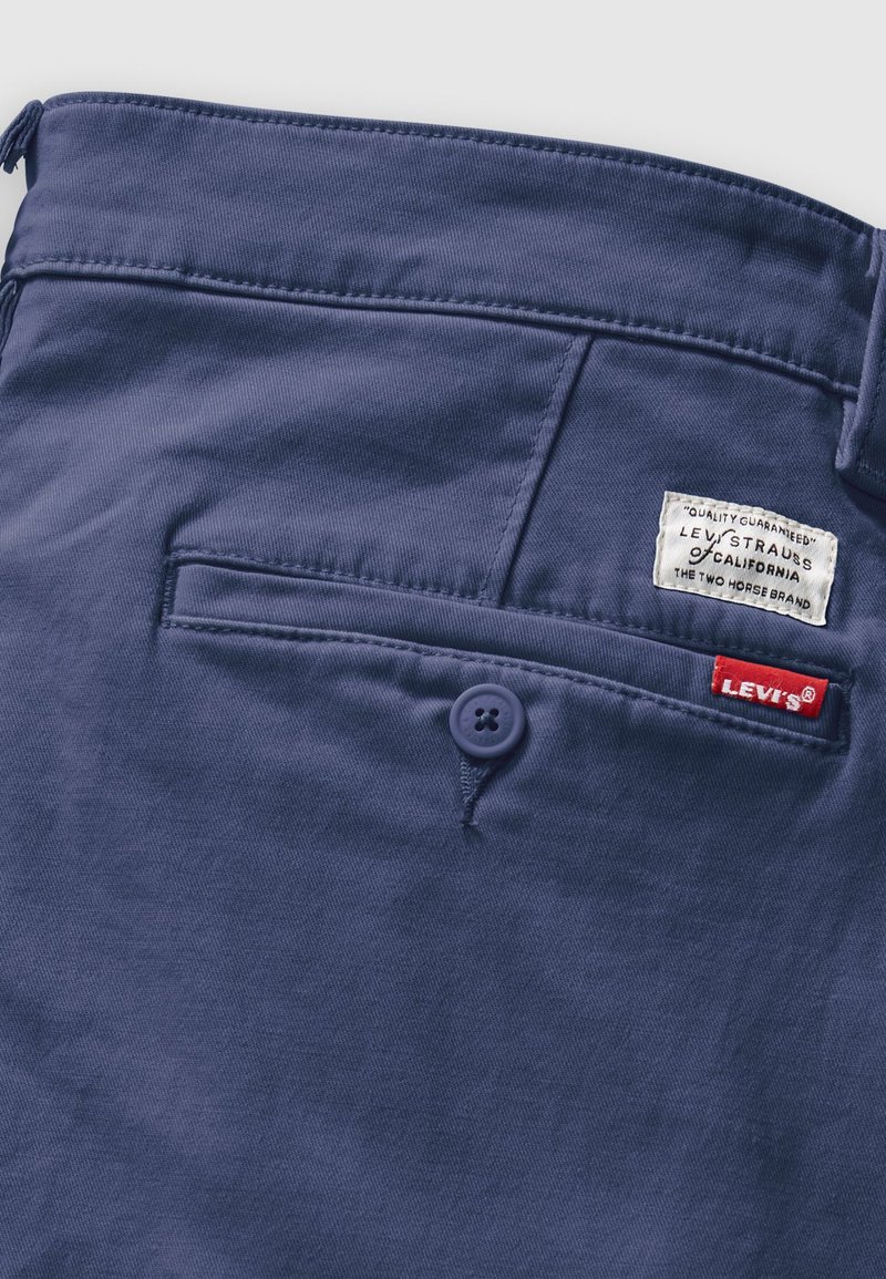 Pantalones azul oscuro con un bolsillo trasero con cierre a botón, una etiqueta blanca con texto y una etiqueta roja de Levi's. Textura de tela suave.