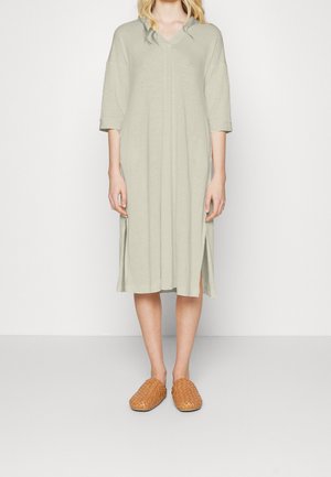 Frau trägt ein locker sitzendes, knielanges beiges Kleid mit dreiviertellangen Ärmeln und braunen gewebten Slip-On-Schuhen, steht vor einem schlichten Hintergrund.