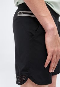 Közeli kép egy fekete rövidnadrágot viselő férfiról, amelyen cipzáras hátsó zseb és szellőző részlet látható az alsó szegélyen, a kéz az oldalon pihen.