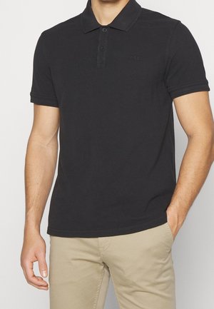 Mies, jolla on musta lyhythihainen poolopaita ja beiget housut, toinen käsi taskussa ja seisoo yksiväristä taustaa vasten.