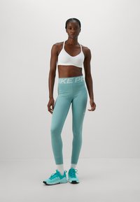 Atletisk kvinde i hvid sportsbh, teal Nike Pro-tights, hvide sokker og teal sneakers, der står mod en ensfarvet grå baggrund.