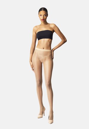 Collants en résille beige avec un motif à pois, associés à un haut bandeau noir. Le mannequin se tient en talons nus contre un fond blanc.