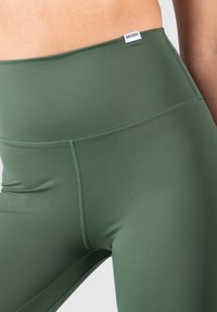 Leggings verdi a vita alta realizzati in un tessuto liscio e elasticizzato. Presentano cuciture minime e un piccolo etichetta del marchio sulla vita.