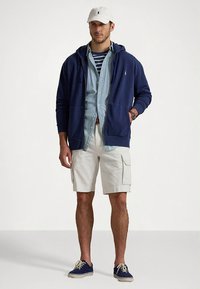 Camisola com capuz azul-marinho, camisa de ganga clara, t-shirt às riscas por baixo, calções cargo bege, boné branco, ténis azuis. Roupa casual em camadas para tempo quente.