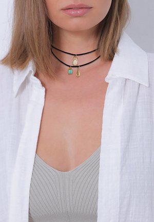 Sort choker med guld accenter og tre vedhæng: en tårnformet, en rektangulær grøn sten og et perleagtigt dråbe. Hvid skjorte med knaplukning synlig.