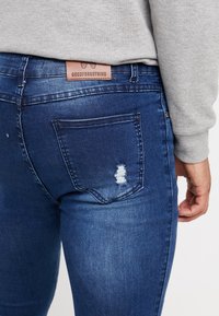 Vaqueros ajustados azules desgastados con un bolsillo trasero desgastado y una etiqueta de cuero marrón con la inscripción "GOODFORNOTHING". Textura de denim suave.