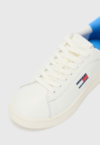 Hvite lær sneakers med rund tå, kanvas snøring og blå innvendig fôr. Har logo-patch i rødt, hvitt og blått på siden.