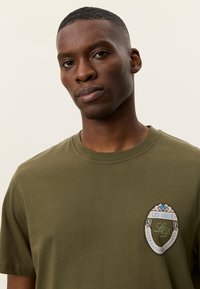 Πράσινο ελιάς βαμβακερό μπλουζάκι με στρογγυλή λαιμόκοψη, κοντά μανίκια, και κυκλικό λογότυπο στο αριστερό στήθος με λουλουδένιες λεπτομέρειες.