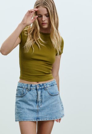 Olivgrünes, eng anliegendes T-Shirt, kombiniert mit einem hellblauen Jeans-Minirock. Der Rock hat einen Knopfverschluss und zwei frontale Taschen.