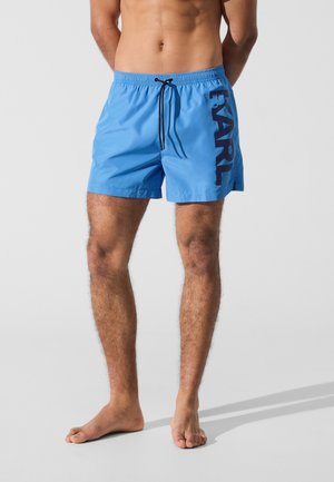 Muž oblečený v modrých plavkových šortkách s čiernym nápisom "KARL" na boku, stojaci bosý na svetlosivom pozadí.