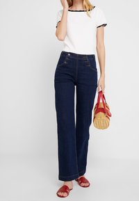 Mujer vestida con una blusa blanca de manga corta y jeans oscuros de tiro alto, sosteniendo un bolso de mimbre con correas rojas y usando sandalias rojas.