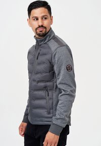 Veste isolée grise avec un design matelassé sur le devant, manches grises texturées, poche zippée et écusson logo sur l'épaule.