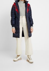 Abrigo tipo puffer marino con acentos en rojo y burdeos oscuro en los hombros, cierre de cremallera y puños acanalados. Combinado con pantalones anchos color crema y botas beige.