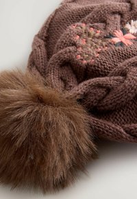 Stickad brun hatt med flätad textur, prydd med blommig broderi i rosa och beige. Inkluderar en stor, fluffig brun pom-pom på toppen.