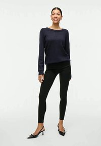 Sweater de malha azul-marinho com mangas longas, leggins pretas e sapatos de salto alto pretos. A modelo está de pé, virada para a frente, a sorrir.