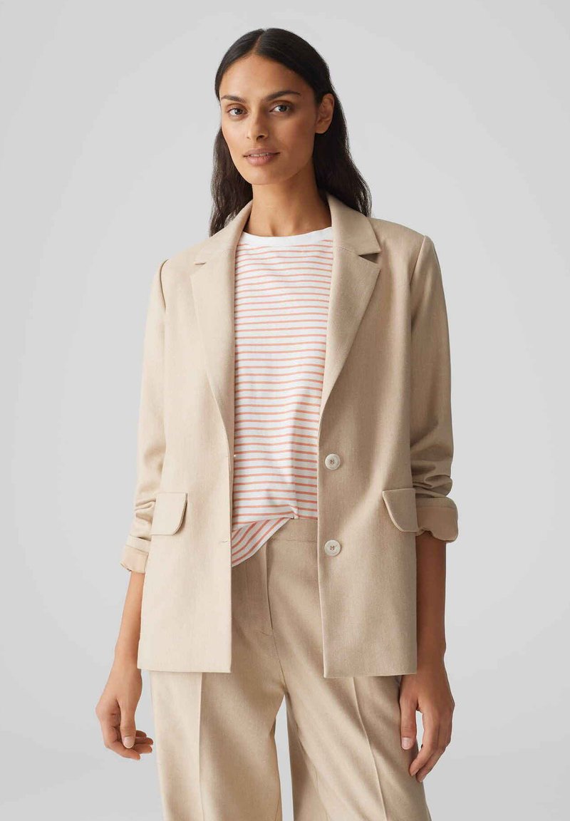 Beige op maat gemaakte blazer met twee knopen aan de voorkant en klepzakken, gedragen over een wit en koraal gestreept overhemd. Eenvoudig, strak ontwerp.