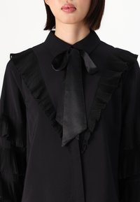 Blusa nera con dettaglio di rouches plissettate lungo il colletto e le maniche, con un fiocco in satin al collo. Tessuto morbido.