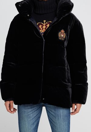 Personne portant une doudoune en velours noir avec un blason brodé sur la poitrine, superposée à un pull en tricot foncé avec un motif de couronne, et un jean bleu.