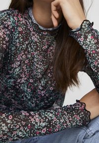 Top lungo a maniche di pizzo nero trasparente con fiori multicolore, bordi ondulati sulle maniche e design aderente.