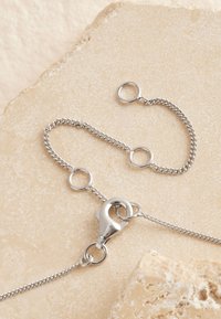 Silberne Metallkette mit Gliedern, versehen mit einem Karabinerverschluss und runden Ringen, vor einer strukturierten Steinoberfläche.