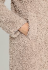 Płaszcz wykonany z jasno-beżowego, pluszowego materiału o miękkiej, teksturowanej powierzchni. Posiada boczną kieszeń i lekko rozkloszowane rękawy.