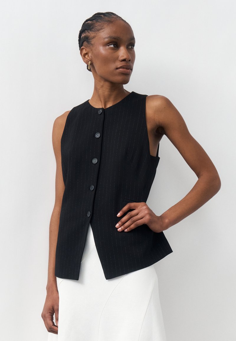 Γυναίκα με πλεξούδες φορεί μαύρο αμάνικο γιλέκο με κουμπιά και λευκή φούστα, ποζάροντας με το χέρι στη μέση της μπροστά από ένα απλό φόντο.