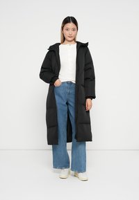 Casaco longo acolchoado preto com fecho de zip e capuz. Usado sobre uma blusa branca e jeans de perna larga em azul claro, combinado com ténis brancos.