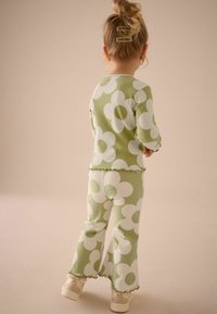 Grünes geripptes Zweiteiler-Outfit mit großen weißen Blumenmustern, ausgestattet mit Rüschenkanten an den Ärmeln und dem Saum, kombiniert mit hellen Schuhen.