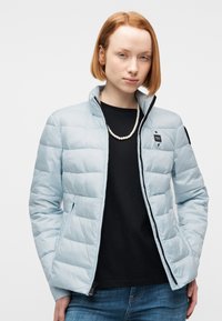 Νεαρός με κοντά κόκκινα μαλλιά φοράει ανοιχτό μπλε καπιτονέ μπουφάν πάνω από μαύρο πουκάμισο και τζιν, συνοδευόμενο από ένα μαργαριταρένιο κολιέ.