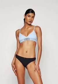 Γυναίκα με σκούρα μαλλιά, ντυμένη με ένα ανοιχτό μπλε bralette Versace και μαύρα μπικίνι, σε στάση μπροστά από ένα ουδέτερο ανοιχτό φόντο.