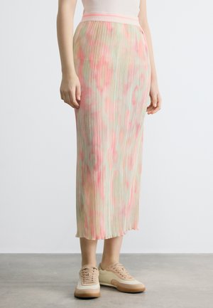 Donna con gonna midi plissettata con motivo astratto rosa e verde abbinata a sneakers beige e bianche, in piedi su un pavimento grigio.