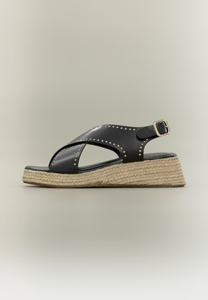 Sandale compensée en cuir noir avec brides croisées, clous dorés, boucle à la cheville et semelle plateforme tressée beige.