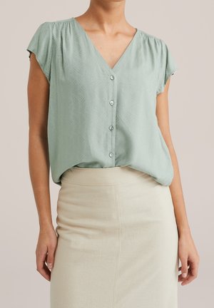 Vrouw draagt een lichtgroene blouse met korte mouwen en knopen, een subtiel patroon en een beige rok met hoge taille.