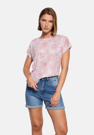 Mujer con una blusa de manga corta estampada con flores rosas y shorts de mezclilla enrollados, de pie con una mano en el bolsillo contra un fondo blanco.