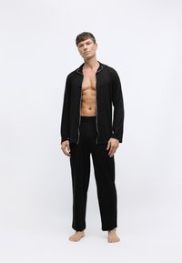 Conjunto de pijama negro de manga larga con ribete blanco contrastante, que incluye una camisa de botones y pantalones con cintura elástica. El modelo va descalzo.