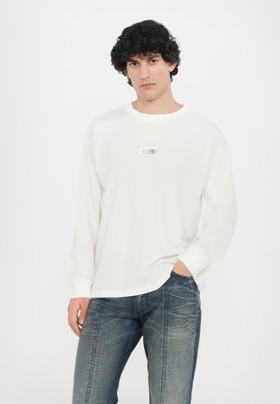 Vit långärmad t-shirt med en liten grafisk design på bröstet, tillsammans med mörkblå, åtsittande denimjeans med texturerade mönster.