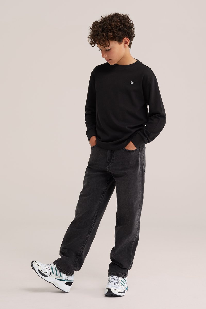 Garçon aux cheveux bouclés portant un t-shirt noir à manches longues, un jean foncé et des baskets blanches, debout les mains dans les poches, regardant vers le bas.