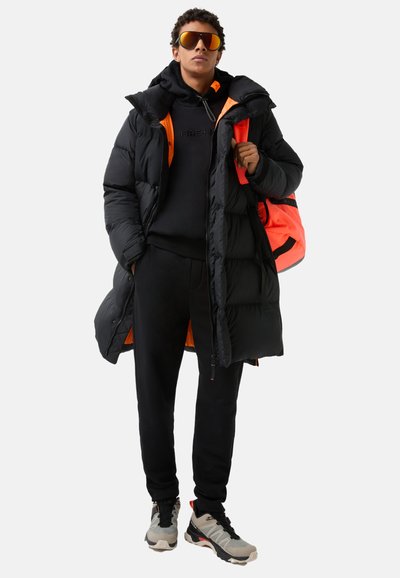 Schwarze Pufferjacke mit orangefarbener Futter, schwarzer Kapuzenpullover, schwarze Jogginghose und graue Sneakers. Das Model trägt einen orange-schwarzen Rucksack.