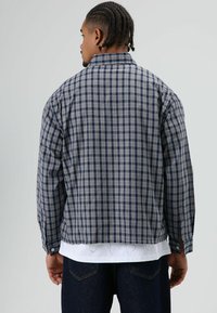 Geruit overhemd in marineblauw en lichtgrijs met een ontspannen pasvorm, knoopmanchetten en een iets kortere snit, gedragen over een wit T-shirt.