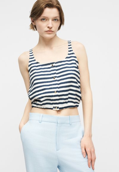 Γυναίκα που φοράει κοντό τοπ χωρίς μανίκια με ναυτικό μπλε και άσπρες ρίγες και κουμπιά, σε συνδυασμό με ανοιχτόχρωμο μπλε παντελόνι, ποζάροντας με το ένα χέρι στην τσέπη.