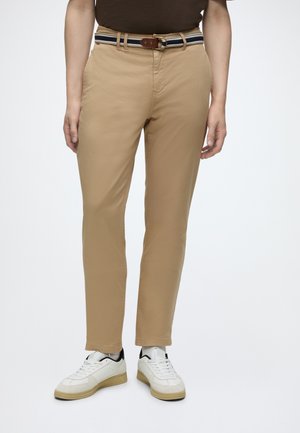 Personne portant un pantalon beige, une ceinture rayée, des baskets blanches avec des semelles beiges, montrée de la taille aux pieds sur un fond clair uni.
