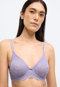 Levandų nėriniuota braletė su banguotu apatine kraštine, reguliuojamomis petnešėlėmis ir maža raktelio formos detalė centre. Tekstūruotas gėlių raštas.