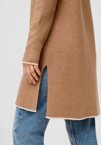 Pull en tricot marron avec une fente sur le côté et des bordures crème, associé à un jean en denim bleu. Le tissu semble doux et texturé.