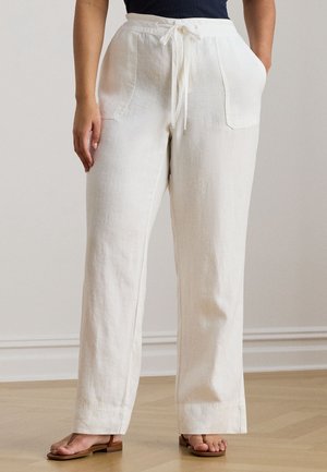 Mujer vestida con pantalones de lino blancos de talle alto con cordón y bolsillos parche, combinados con sandalias planas marrones, de pie en el interior sobre suelo de madera.