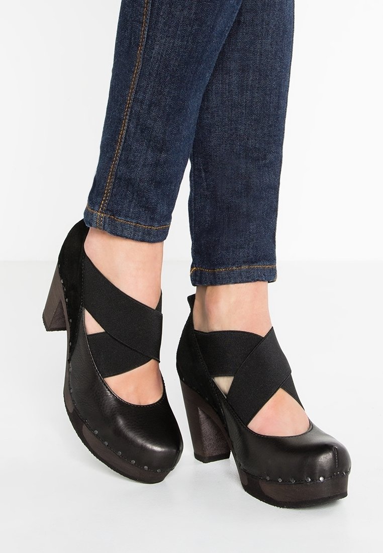 Chaussures à talons noires avec brides croisées élastiques, dessus en cuir texturé, bout rond et clous décoratifs argentés le long de la semelle.