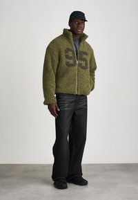 Giacca in pile verde con chiusura a zip e grande grafica nera "35" sul davanti, abbinata a jeans neri a gamba larga e un cappellino.