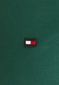 Zelená tkanina s texturovaným povrchem, na které se nachází malá vyšitá nášivka s logem: černý obdélník se červenými, bílými a modrými prvky uvnitř.