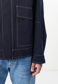 Mornarska jakna z belimi šivi, z majhnim žepom na prsih in sproščenim krojem. V kombinaciji s svetlo modrimi kavbojkami, ki poudarjajo vsakodnevno obleko.